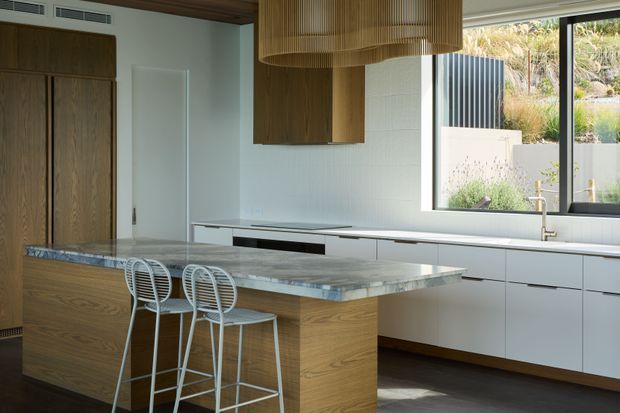 A refined contemporary kitchen with a large island clad in warm timber cabinetry and topped with a grey veined stone benchtop. White flat-panel wall cabinetry and integrated appliances line the rear wall, while two slim white metal bar stools sit at the island. A sculptural timber slatted pendant hangs above, with large black-framed windows bringing in natural light and views of landscaped planting. The space has a clean, warm, understated character.