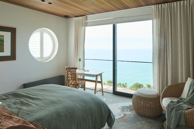 A serene coastal bedroom with a low bed, muted green bedding, a compact timber desk and chair, woven rattan seating, and full-height glazed doors opening to a balcony with ocean views. The room features a warm timber slatted ceiling, pale walls, textured curtains, a distinctive round porthole-style window, and soft natural light.