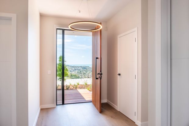 A bright contemporary residential entryway with pale walls, light timber flooring, a tall glazed door opening onto a timber deck, and a sculptural circular pendant light. Built-in white cabinetry and a flush interior door line the hallway, while a landscaped outdoor terrace and distant view are visible beyond the entry.