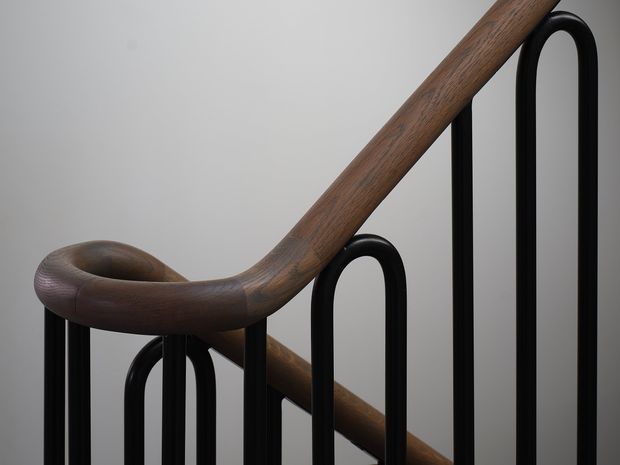 Close-up detail of a sculptural staircase handrail, showing a smoothly curved dark timber rail paired with slender matte-black metal balusters against a plain light-grey background. The image focuses on the craftsmanship, organic curve, and contrast between wood and metal.