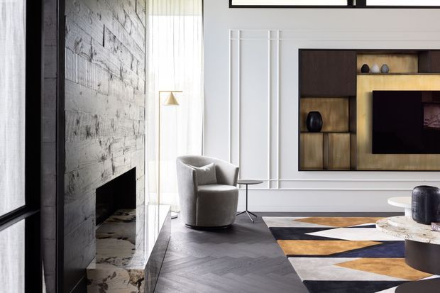 Refined contemporary living room with a dark chevron timber floor, a textured pale stone fireplace wall, white decorative wall panelling, and a bespoke dark timber and brass-toned media cabinet. The room includes a curved upholstered armchair, sculptural side table, geometric area rug, and large glazed opening with sheer curtains.