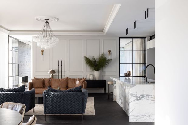 Refined open-plan living and kitchen interior with white decorative wall panelling, dark timber flooring, a marble waterfall island, black-framed glazed doors, and sculptural contemporary furniture. The living area includes a tan sofa, navy quilted armchairs, a patterned rug, and a statement globe chandelier, creating a sophisticated modern-classic aesthetic.
