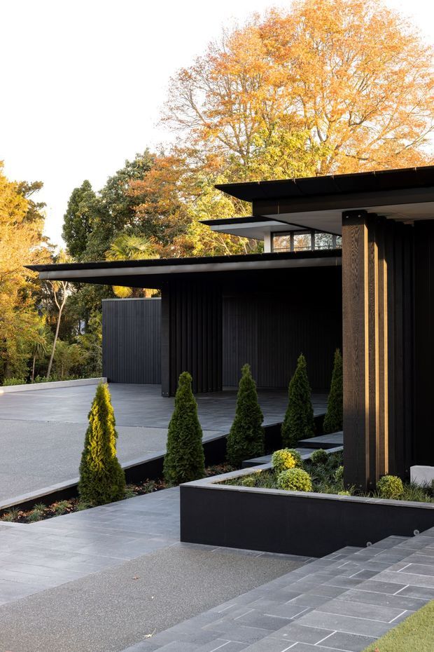Contemporary dark-clad residential exterior with strongly articulated flat roof planes, deep overhangs, and vertical ribbed wall cladding. The building opens onto a carefully landscaped courtyard with layered grey paving, raised black planter beds, clipped conifers, and mature deciduous trees in autumn colour.