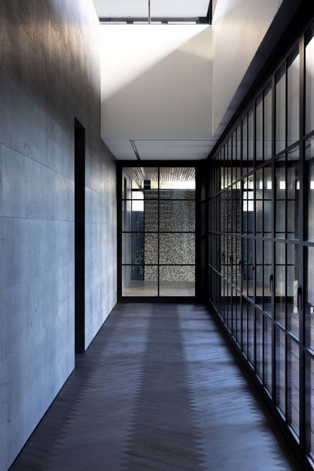 A narrow, dramatic interior passage with a dark zigzag-patterned timber floor, large grey concrete wall panels on the left, and a full-height black-framed glazed wall on the right. A skylight and clerestory glazing bring in strong natural light, while a glazed opening at the end reveals a textured masonry screen and planted courtyard beyond.