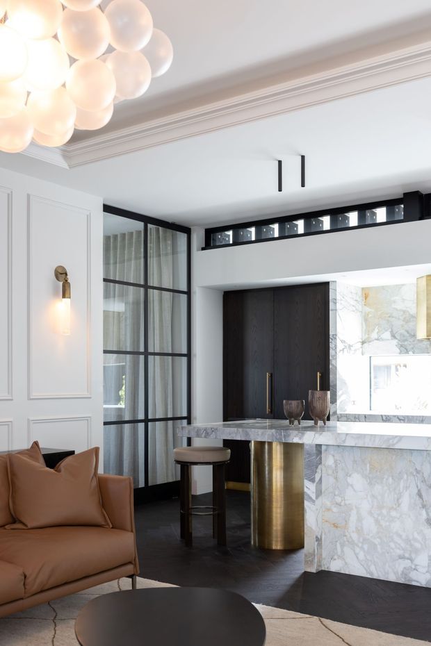 A refined open-plan interior combining a living area and compact kitchen or bar. The room features pale grey decorative wall panelling with ornate cornice detailing, black-framed internal glazing, dark timber cabinetry, a veined marble island with cylindrical brass-gold supports, and a sculptural cluster pendant light. A caramel leather armchair and dark round coffee table add warmth to the sophisticated scheme.