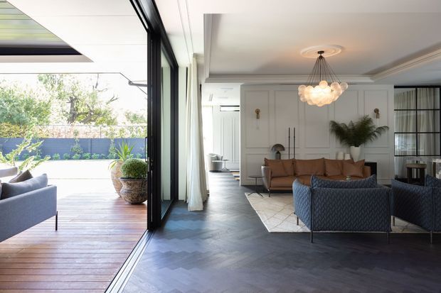 A refined contemporary living room opening directly onto a timber deck through fully retractable black-framed glazing. The interior features dark herringbone timber flooring, a white decorative wall-panelled fireplace or media wall, tailored sofas and armchairs, a sculptural cluster pendant light, and neutral furnishings. The adjoining outdoor area includes built-in seating and potted greenery.