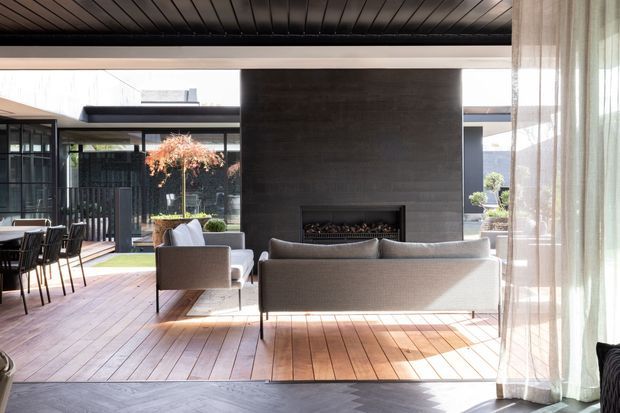 A refined open-plan living room with a dining area, expansive glazing, and a central dark concrete fireplace wall. The room features timber flooring, a dark timber-lined ceiling, neutral upholstered sofas and armchairs, black-framed glass doors, and sheer curtains opening toward a landscaped courtyard.