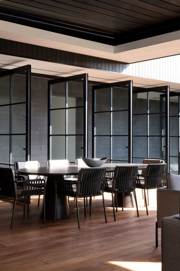 A refined dining area with a long dark dining table surrounded by black woven-back chairs. The room features warm timber flooring, dark framed glazed partition doors, a grey stone or concrete-look wall, and a striking black slatted ceiling with clerestory-style glazing that brings in strong natural light.