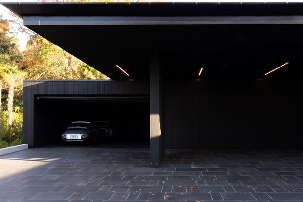 A dark, minimalist modern carport and garage exterior with a broad flat roof, black vertical cladding, recessed linear lighting, a central structural post, and a paved forecourt. One vehicle is visible in the enclosed garage bay, with landscaped trees at the edge of the frame.