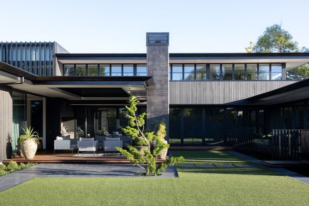 Contemporary multi-wing residence with dark metal framing, extensive floor-to-ceiling glazing, horizontal rooflines, timber-clad upper walls, and a tall central masonry chimney. The home opens onto a landscaped courtyard with a covered outdoor lounge, paved terrace, ornamental planting, and lawn.