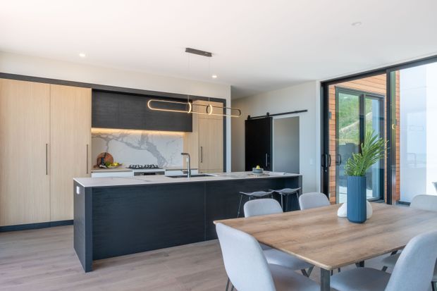 Bright open-plan kitchen and dining area with full-height light wood cabinetry, a dark kitchen island, marble-look splashback, integrated appliances, and sculptural linear pendant lighting. A timber dining table with upholstered chairs occupies the foreground, while large glazed doors and timber exterior cladding are visible at right.