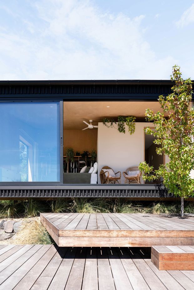 A contemporary single-storey home viewed from a timber deck, with a dark corrugated exterior frame, expansive sliding glazing, and a covered indoor-outdoor living area. The recessed patio contains wicker lounge chairs, a white ceiling fan, planted greenery, and a small dining or kitchen area visible inside. Layered timber decking and raised platforms occupy the foreground, with native-style grasses and a tree integrated into the landscaping.