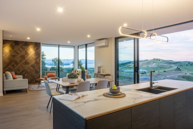 A contemporary open-plan kitchen and dining area with a large marble-look island, dark wood cabinetry, and sculptural pendant lighting. Floor-to-ceiling glazing opens to expansive rolling green hills and a water view. A small living area with a geometric timber feature wall is visible in the background.