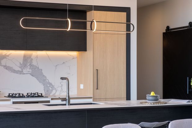 Contemporary kitchen featuring dark timber cabinetry, light timber pantry cabinetry, a white marble-look splashback and benchtop, a central island with integrated sink, and a sculptural linear pendant light. A dark sliding door and partial dining seating are visible beyond the island.