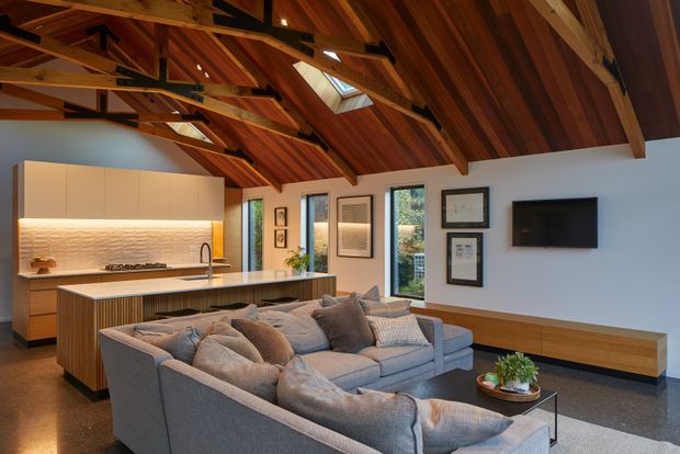 Open-plan living room with a contemporary kitchen, large island, and generous sectional sofa beneath a dramatic exposed timber vaulted ceiling. The ceiling incorporates dark steel structural connectors and a skylight. Warm timber ceiling lining and cabinetry contrast with pale walls, a stone-toned island benchtop, dark flooring, framed artwork, and a wall-mounted television.
