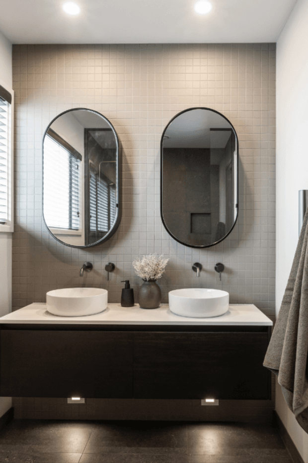 A contemporary bathroom featuring a double floating vanity with two white vessel basins, dark wood cabinetry, a pale stone or solid-surface benchtop, and a full-height small square tiled feature wall. Two black-framed arched mirrors sit above wall-mounted black tapware. Dark floor tiles, recessed ceiling lights, and a window with blinds complete the refined, warm-toned interior.