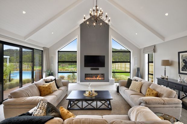 Spacious, bright living room with a dramatic vaulted white ceiling and exposed beams, symmetrical gable-end glazing, black-framed sliding doors, and views to a pool and landscaped grounds. Two large neutral sofas and additional seating surround a dark rectangular coffee table, while a central charcoal feature wall incorporates a television and fireplace. A substantial chandelier provides a decorative focal point.