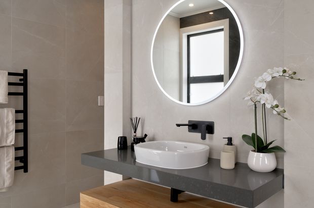 Contemporary bathroom vanity with a floating dark grey stone benchtop, white vessel basin, black wall-mounted tapware, round illuminated mirror, large light-grey wall tiles, timber open shelving, and a black heated towel rail. A potted orchid and coordinated accessories add a refined natural accent.
