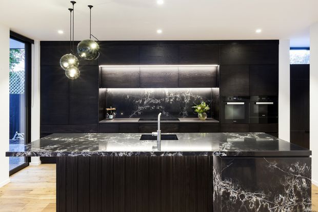 Contemporary kitchen featuring a large dark marble island with waterfall end panel, black timber-look cabinetry, integrated appliances, illuminated shelving, and spherical pendant lights. Light timber flooring and outdoor views through glazing provide contrast to the dramatic dark finishes.