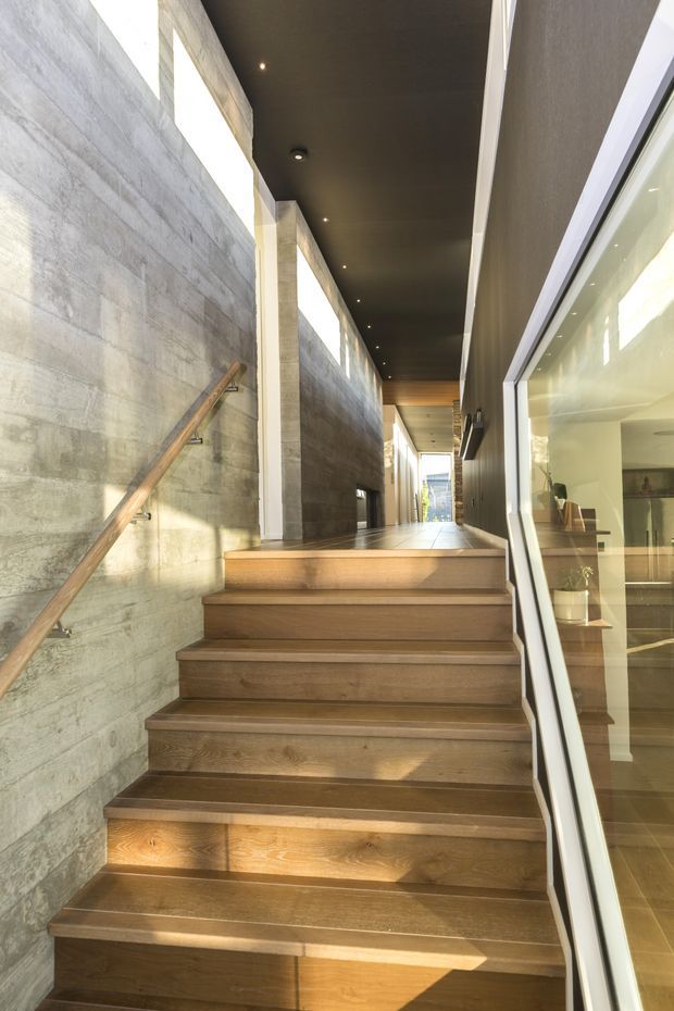 A modern interior stairway rises through a long, narrow corridor. Wide timber stairs and a timber handrail contrast with textured grey concrete walls, while a dark ceiling with recessed downlights and tall clerestory glazing create a dramatic, linear composition. Full-height glazing on the right reveals built-in interior joinery and a bright adjoining space.