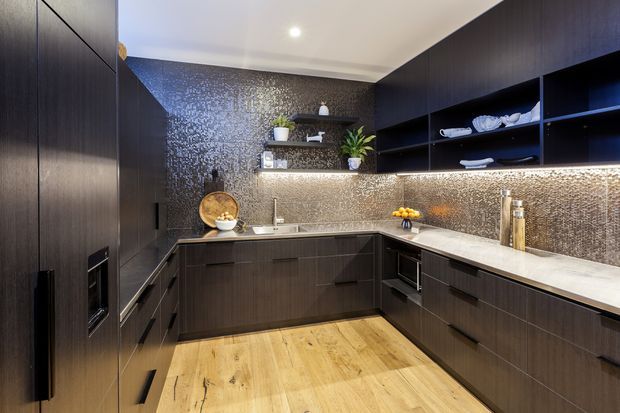 A sophisticated dark-toned contemporary kitchen with a U-shaped layout, tall integrated cabinetry, open shelving, a pale stone benchtop, textured metallic splashback, and warm timber flooring. Under-cabinet lighting highlights the reflective splashback and creates a refined, dramatic atmosphere.