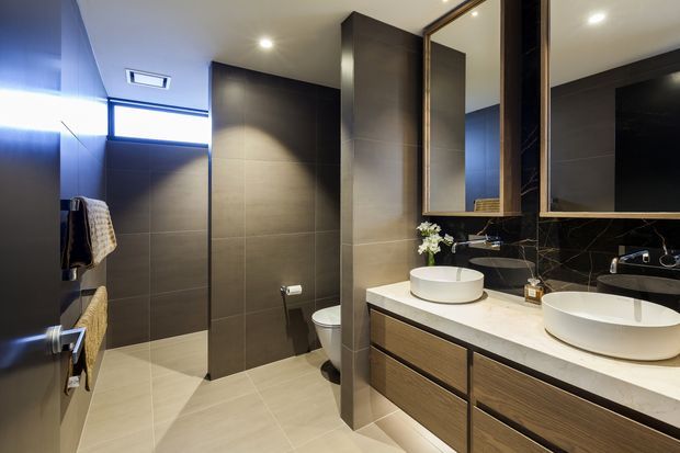 Contemporary bathroom with a spacious double-vanity, two vessel basins on a light stone benchtop, dark timber cabinetry, large-format grey wall and floor tiles, black marble-look splashback, mirrored cabinetry, a toilet, and a partially enclosed shower area. Recessed ceiling lighting and a high horizontal window provide a refined, softly illuminated interior.