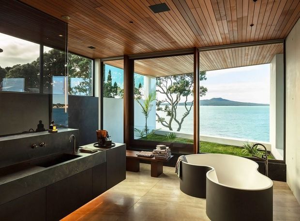 A refined modern bathroom with a freestanding sculptural bathtub positioned beside expansive floor-to-ceiling glazing overlooking turquoise water and a distant mountainous shoreline. The room features dark built-in vanity cabinetry with an integrated basin, warm timber slatted ceiling, large tiled flooring, dark timber window and door frames, and a small indoor plant. The strong indoor-outdoor connection and waterfront outlook create a calm, luxurious atmosphere.