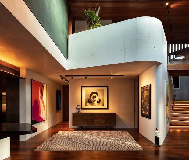 A dramatic contemporary residential interior featuring a double-height entry and living area, curved white mezzanine balustrade, warm timber ceiling and flooring, sculptural staircase, recessed artwork display, dark timber sideboard, and a large patterned rug. Track lighting and warm indirect illumination create a refined, gallery-like atmosphere.