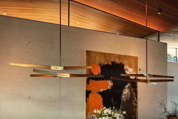 Interior view of a refined contemporary space with a warm slatted timber ceiling, smooth neutral plaster walls, and dramatic suspended linear pendant lighting. A large abstract artwork forms a bold focal point, with floral arrangements visible below. Glazing and a partial adjoining area are visible at the right.