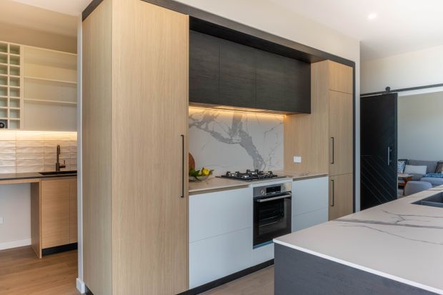 Contemporary kitchen with a compact built-in cooking zone, pale timber cabinetry, dark upper shelving and trim, white lower cabinets, and a dramatic marble-look splashback and island benchtop. A pantry or utility area with open shelving is visible to the left, while a living area is visible through the dark doorway on the right.