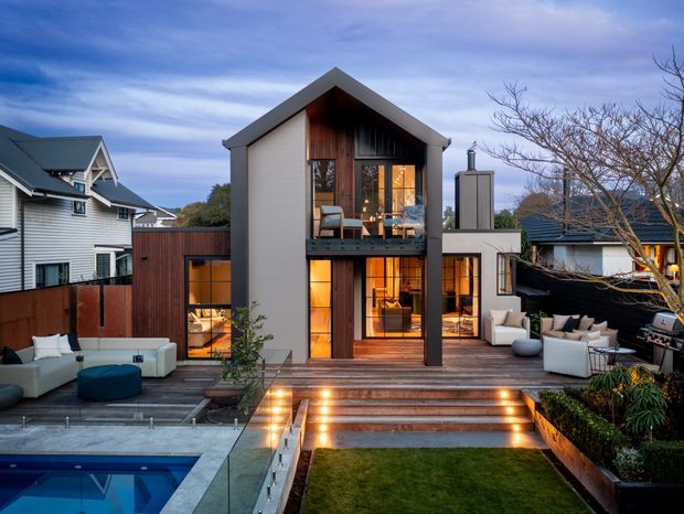 Contemporary multi-level residential home viewed from the rear, featuring a steep gabled roof, white rendered walls, extensive warm timber-framed glazing, and a large timber deck with illuminated steps. The outdoor entertaining area includes lounge seating, built-in planting, a lawn, and a swimming pool in the foreground. The surrounding suburban setting and neighbouring homes are visible.