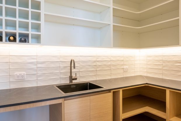 A compact contemporary kitchen with an L-shaped dark benchtop, integrated black sink and brushed metal tap, light timber cabinetry, open shelving, and white textured rectangular tiled splashback. The cabinetry and shelving are painted white above the work surface, creating a clean, bright fit-out.