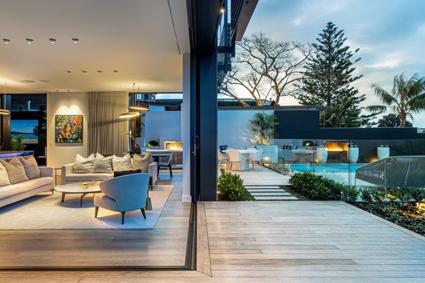 A contemporary indoor-outdoor living space opening onto a timber deck and landscaped pool terrace. The interior features a neutral sectional sofa, lounge chair, rug, artwork and modern lighting, while the exterior includes a glass pool fence, outdoor dining area, dark privacy wall and tropical planting.