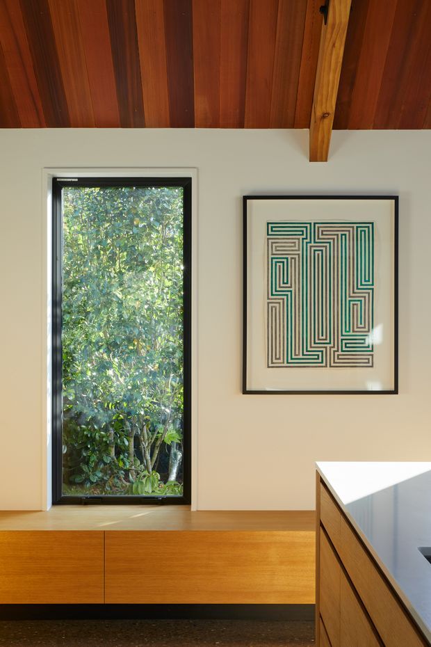 A warm, refined interior detail showing a compact kitchen or built-in joinery area. Vertical timber-lined ceiling boards and an exposed timber beam frame a pale plaster wall with a tall black-framed window overlooking leafy vegetation. A framed geometric artwork hangs beside the window. Below, a long timber built-in cabinet or bench runs along the wall, while the foreground shows a timber kitchen island with a light stone benchtop. The image has a strong connection to natural materials and greenery, with clean modern detailing.