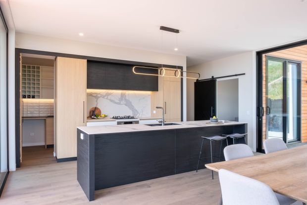 A bright contemporary open-plan kitchen and dining area with a large dark island, pale timber cabinetry, stone benchtops and splashback, integrated wine storage, and minimalist pendant lighting. Full-height glazing opens toward an exterior timber-clad area, while a timber dining table and upholstered chairs occupy the foreground.