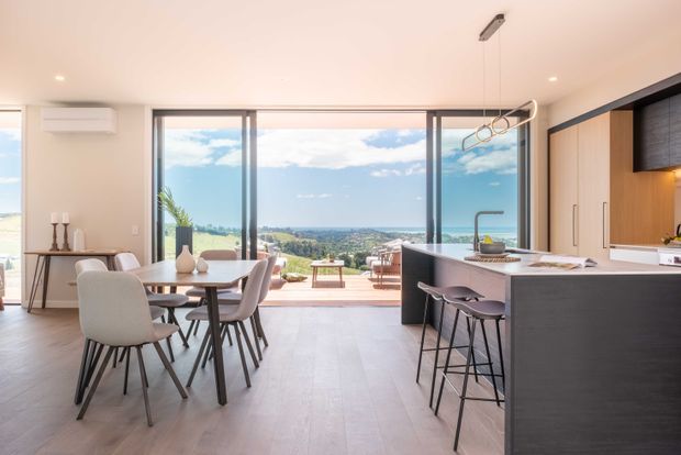 Bright open-plan kitchen and dining area with a large island, integrated cabinetry, pendant-style hanging rack, and dining table. Full-height glazing opens to a terrace and expansive coastal or suburban views. The interior uses pale timber-look flooring, light walls, dark cabinetry, and contemporary furnishings.