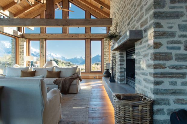 A spacious rustic living room with a double-height timber ceiling and exposed structural beams, large timber-framed windows overlooking dramatic snow-capped mountains, and a substantial rough stone fireplace. Soft upholstered sofas and armchairs, a pale rug, and warm timber flooring create a comfortable alpine retreat atmosphere.