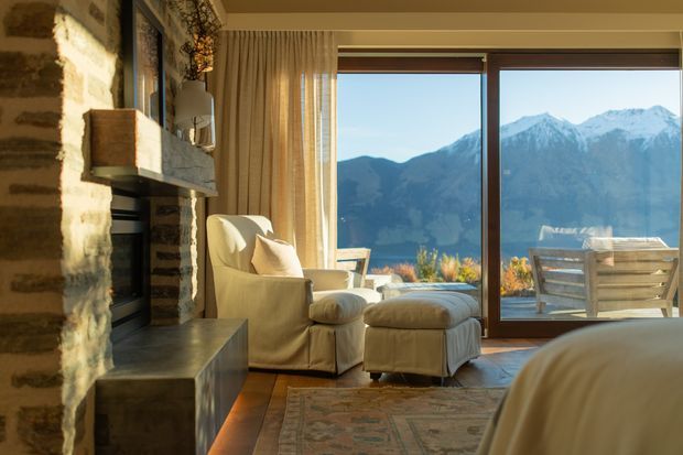 Warm mountain-view living space with a rustic stone fireplace wall, large timber-framed glazing, a cream upholstered armchair and ottoman, and a partial bed visible in the foreground. The room opens visually toward a balcony or terrace overlooking snow-capped mountains. Natural timber flooring, woven curtains, and earthy textiles create a cozy alpine retreat atmosphere.