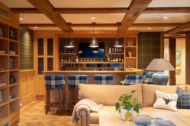 A warm, richly detailed home bar and lounge area. The room features extensive timber cabinetry and shelving, exposed dark timber ceiling beams, a built-in bar with seating for several people, pendant lights, a television, bottle and glass storage, and a cream upholstered sofa in the foreground. Timber parquet flooring and patterned wall coverings add texture and a traditional lodge-like character.