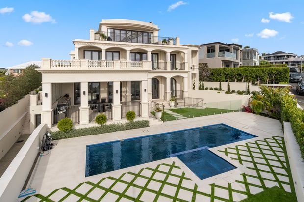 Exterior view of a large multi-storey villa with a classical Mediterranean-inspired facade, arched openings, columns, balustraded terraces and extensive glazing. The property includes a rectangular swimming pool, landscaped lawn with geometric paving, perimeter walls and hedging, with neighbouring homes visible beyond.