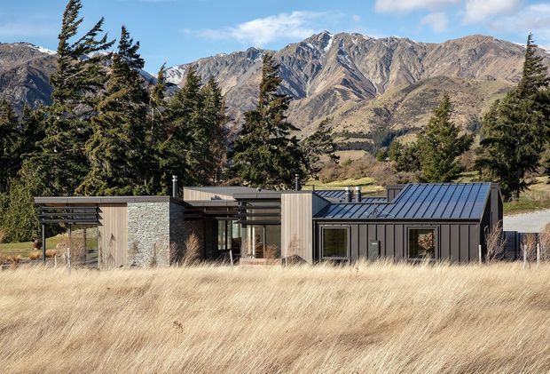 A low-profile contemporary house set in a dry alpine landscape, with rugged mountain ranges and tall evergreen trees behind it. The building combines dark vertical metal cladding, pale timber-clad sections, and a textured stone wall, with broad horizontal roof planes and a pitched metal roof over the right-hand volume. Long horizontal glazing and covered outdoor areas create a strong connection to the surrounding grassland.