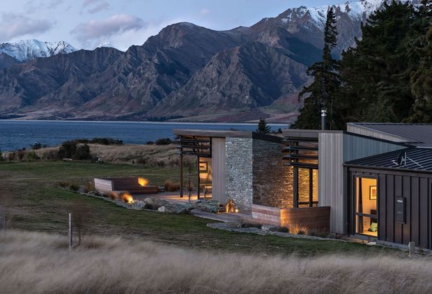 A modern single-storey pavilion-style house set beside a lake, with dramatic snow-capped mountain scenery behind it. The building features dark metal cladding and roofing, vertical timber-look screens, extensive glazing, and a prominent natural stone feature wall. Warm interior lighting and small outdoor fire features create a cosy evening atmosphere, while the surrounding landscape is open and rural.