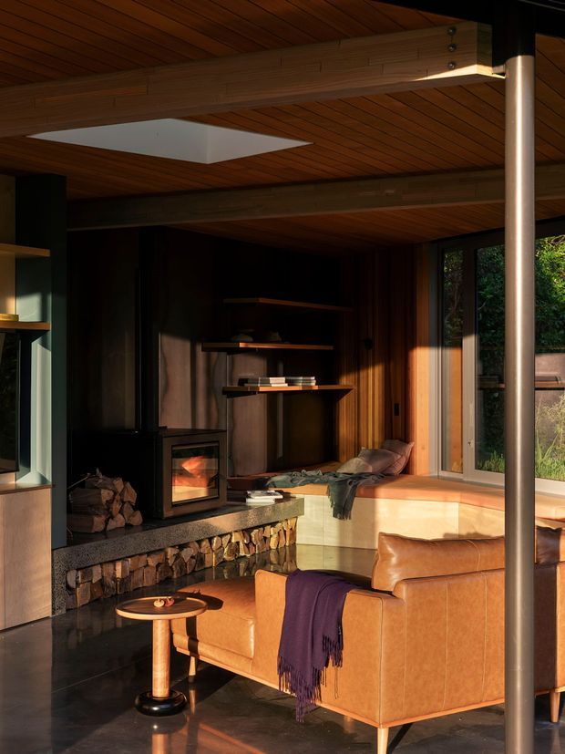 Warm, moody living room with a wood-lined ceiling, exposed structural beams, dark timber wall panelling, and a stone-clad fireplace with a wood-burning insert. A leather sofa and lounge chair face the fireplace, while large windows bring in strong afternoon light and views of greenery. A roof skylight adds daylight above the seating area.