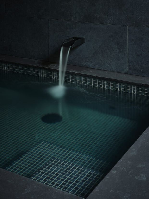 Close-up of a dark, moody plunge pool or spa with a wall-mounted angular black water spout pouring into the pool. The pool interior is finished with small dark teal square mosaic tiles, while the surrounding coping and wall are clad in large charcoal stone-look tiles.