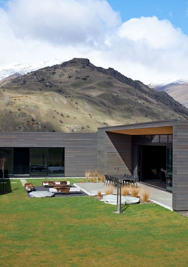 A low, modern single-storey house arranged around a landscaped courtyard, set against a dramatic mountainous alpine landscape. The building has dark horizontal timber cladding, expansive black-framed glazing, and a deep recessed entry or outdoor entertaining area with a timber-lined soffit. A manicured lawn, paved terrace, outdoor dining furniture, sculptural fire-pit seating, ornamental grasses, and large stepping stones occupy the foreground.