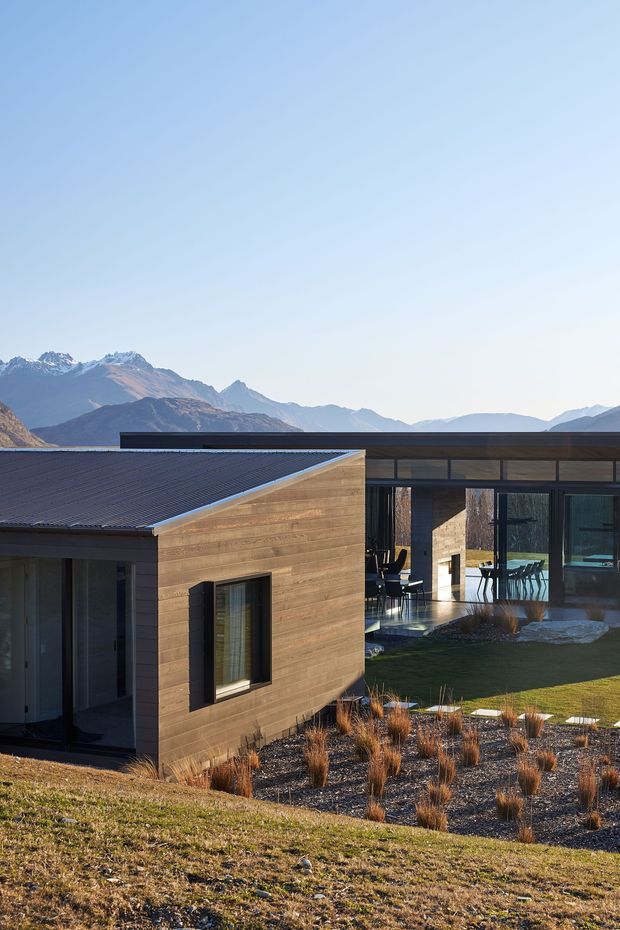 Exterior view of a contemporary single-storey home set on a landscaped sloping site, with warm horizontal timber-clad walls, dark low-pitched roofing, extensive glazed openings, and a covered outdoor living area. Mountain ranges are visible in the background.