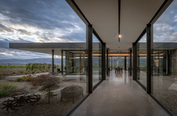 A dramatic modern residential passage or entry gallery with a long polished concrete floor, extensive floor-to-ceiling glazing, dark slender structural frames, and a simple flat roof. The glazed corridor connects to a dining area visible at the far end and opens onto a sparse gravel landscape with sculptural fire features and broad mountain views. The architecture emphasizes transparency, strong horizontal and vertical lines, and indoor-outdoor connection.