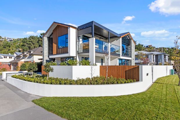 Exterior view of a contemporary multi-level residence with asymmetrical pitched roof forms, dark metal framing, timber cladding, white rendered walls, expansive glazing, and a covered upper-level balcony. The home is set behind a curved white boundary wall and landscaped front garden in a suburban neighbourhood.