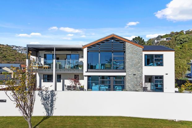 Contemporary multi-level residential home viewed from the street, with white rendered walls, a central gabled stone-clad feature wall, extensive blue-tinted glazing, dark horizontal louvres, and balconies. The house sits on a landscaped sloping suburban site with hillside homes visible in the background.