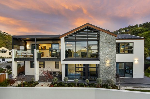 Exterior view of a contemporary multi-level residential house at dusk. The facade combines light rendered walls, a prominent natural stone-clad gable volume, dark metal-framed glazing, balconies, and a mixed pitched and low-pitched roof form. Landscaped planting and neighbouring hillside homes are visible around the property.